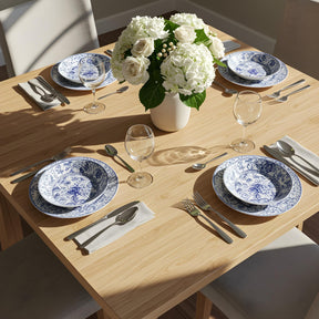 Close-up of a light wood dining table refinished using Retique It Pickled Oak grained kit, styled with white and blue floral china, clear wine glasses, and a centerpiece of white roses and hydrangeas. The soft faux oak finish adds a refined, natural look perfect for any home dining space.