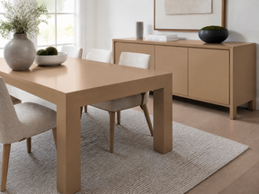 A minimalist dining room scene featuring a light oak rectangular dining table with chunky block legs, surrounded by neutral upholstered chairs, set on a textured beige rug. In the background, a matching light oak sideboard cabinet is styled with decorative objects.